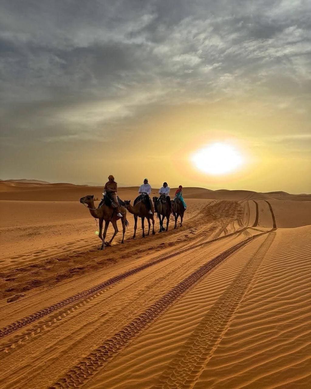 Camel riders crossing sand dunes at sunset